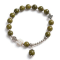 A beaded stretch bracelet featuring olive green stone beads, silver-toned spacer beads, a white carved accent bead, a dangling green bead charm, and silver decorative charms, shown on a white background.