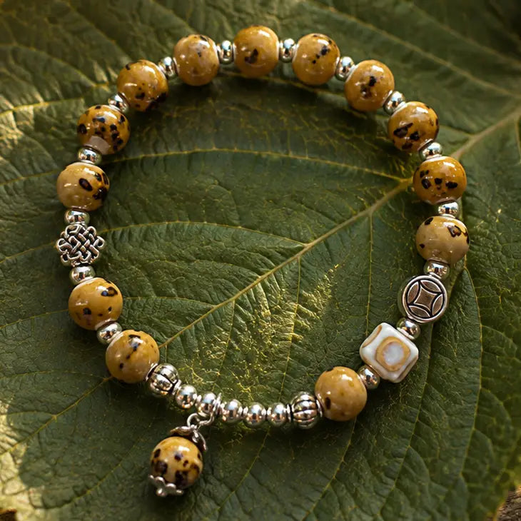 A beaded bracelet with round tan and black marbled beads, silver-toned spacer beads, a silver knot charm, a white square bead with a brown center, and a dangling marbled bead charm, displayed on a green leaf.