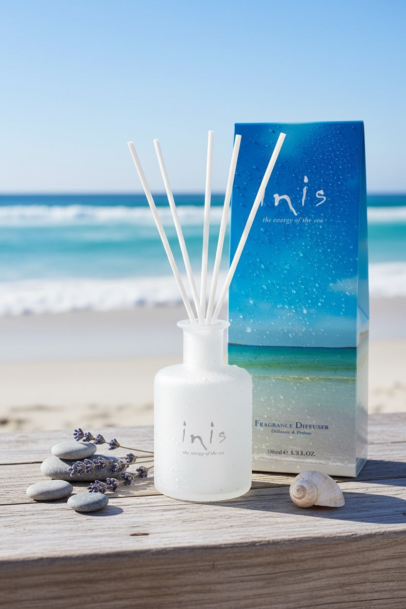 Inis Fragrance Diffuser bottle and box on driftwood at the beach with lavender sprigs, pebbles and a seashell